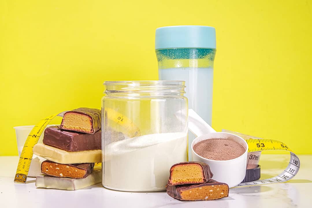 Protein bars, nutrition drink, supplement powder, and measuring tape placed in a table with a yellow background.